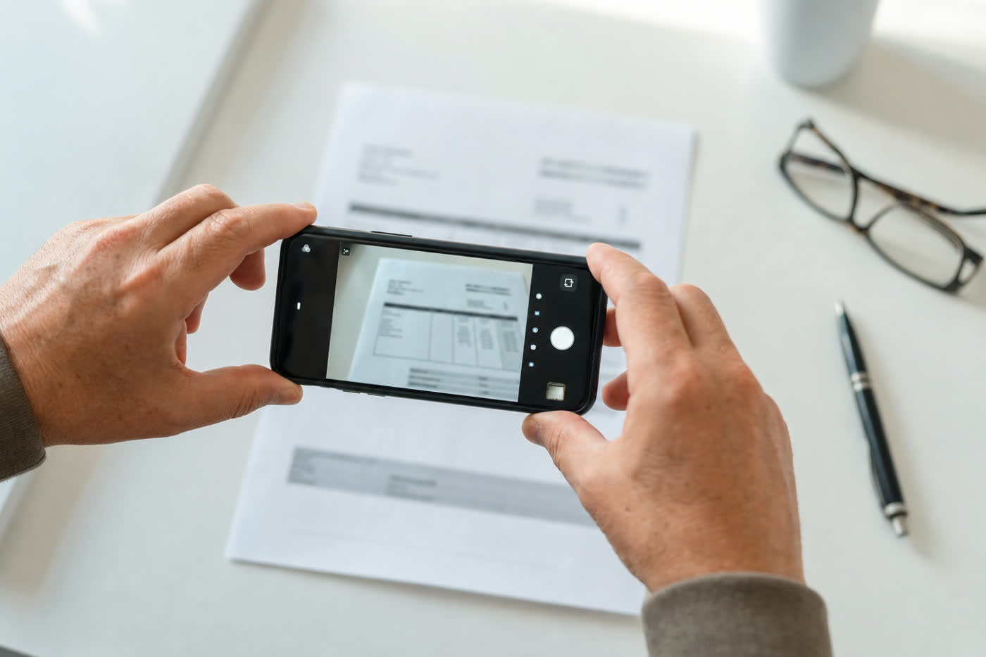 Zwei Hände fotografieren eine Papierrechnung mit dem Smartphone am Schreibtisch
