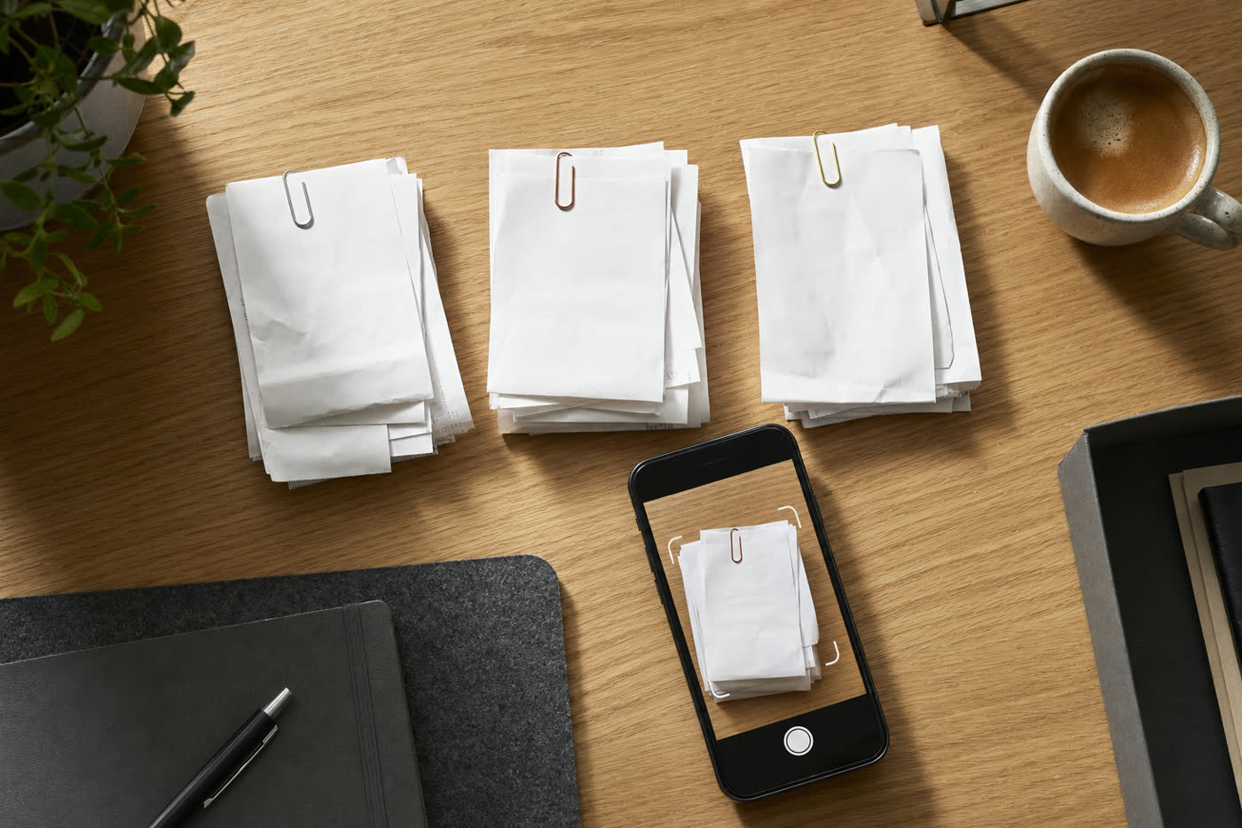 Aufgeräumter Schreibtisch: sortierte Belegstapel mit Büroklammern, daneben ein Smartphone beim Abfotografieren eines Kassenbons